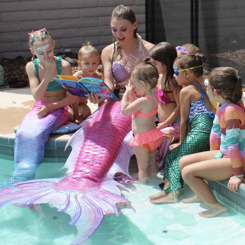 Photo of a woman dressed as a mermaid with a bright pink tail reading a book to a group of children