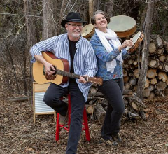 man with a guitar and woman with a drum infront of a wooded area