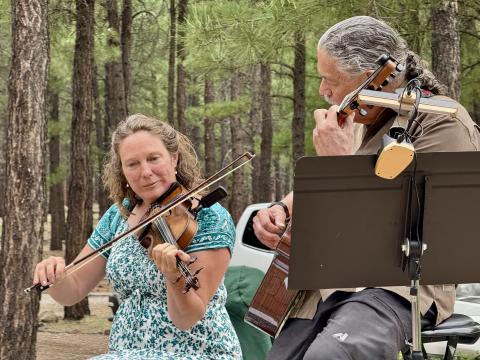 woman playing violin, man playing guitar against background of trees