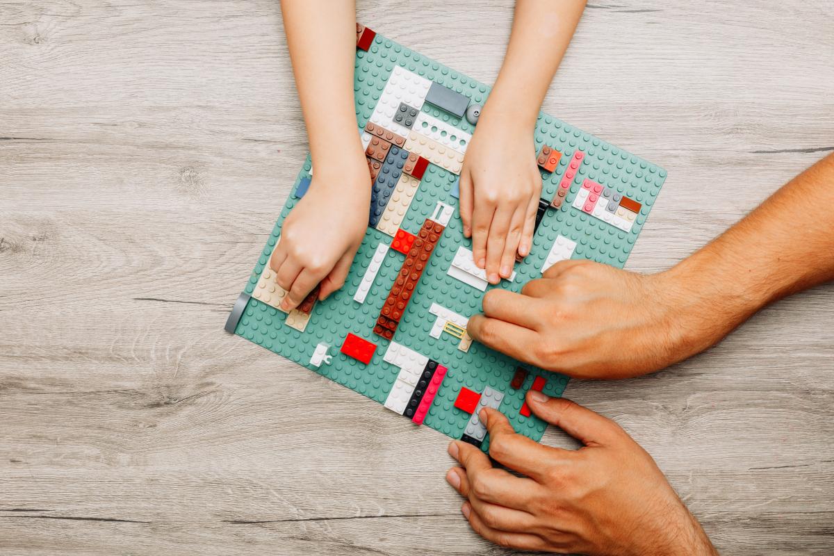 two paris of hands, one adult and one child, attaching colorful Lego pieces to a green pegboard