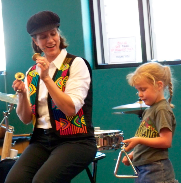 woman in a hat and colorful vest showing a small girl how to play a triangle